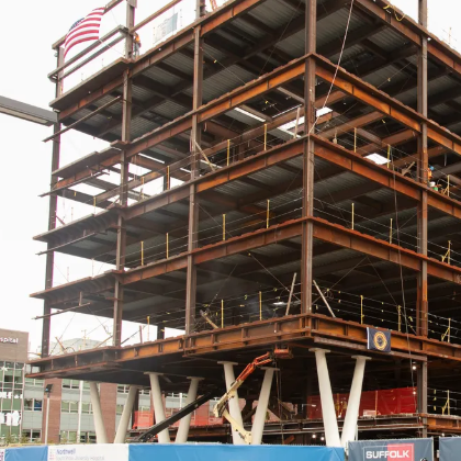 A multi-story steel building framework under construction, with cranes and construction equipment nearby. Blue fencing and signs, including Northwell South Shore University Hospital, are visible at the site.