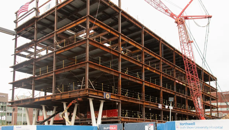 A multi-story steel building framework under construction, with cranes and construction equipment nearby. Blue fencing and signs, including Northwell South Shore University Hospital, are visible at the site.