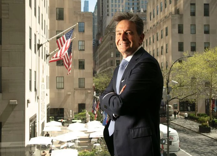 A man in a dark suit stands with arms crossed, smiling in front of a window overlooking a city street with American flags, tall buildings, and outdoor seating below.