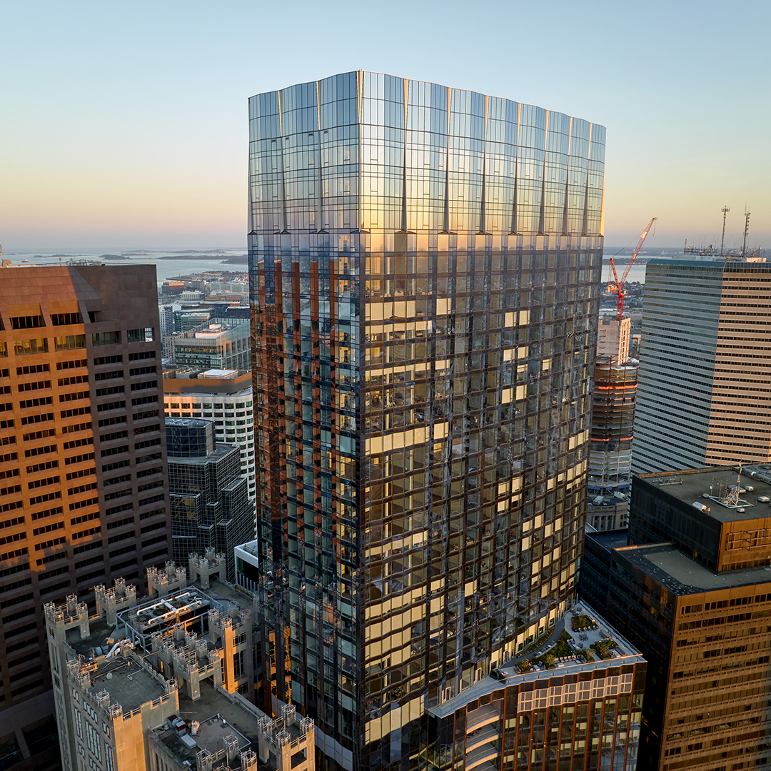 A modern glass skyscraper reflects the evening light in a city skyline, surrounded by other tall office buildings and cranes, with a distant view of water under a clear sky.