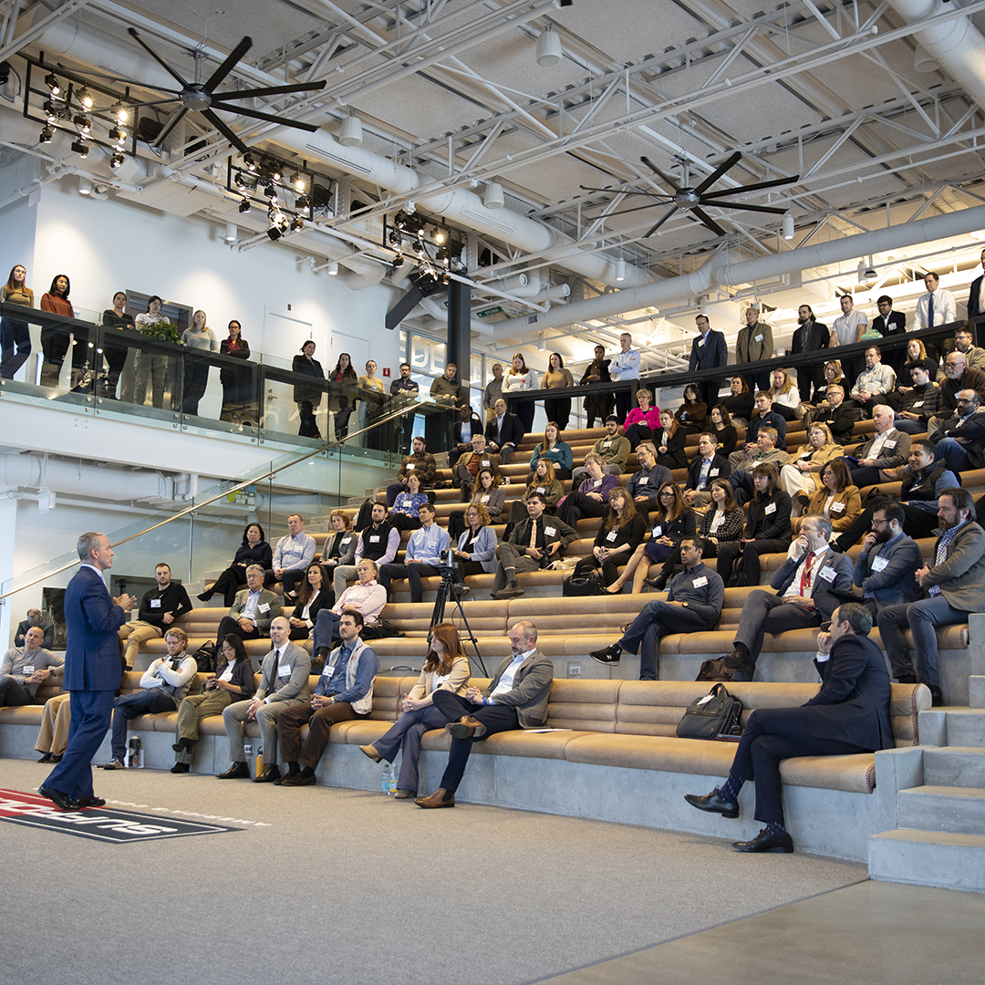 A man in a suit speaks to a large audience seated on tiered steps in a modern, open space with glass railings, high ceilings, and industrial lighting. People listen attentively, some seated above on a mezzanine.