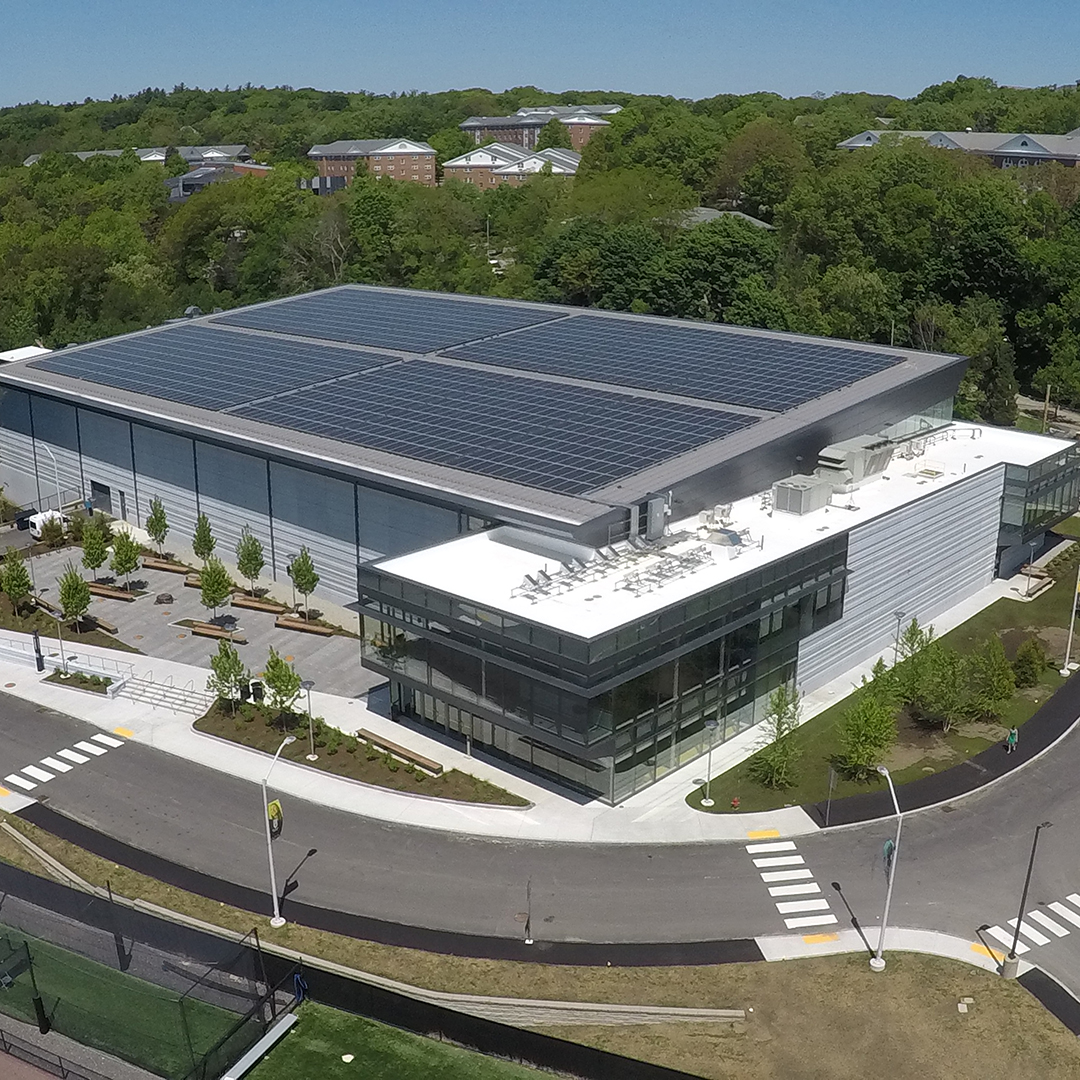 A modern building with large solar panels on the roof, surrounded by trees and greenery. Wide walkways, landscaped areas, and parking spaces are visible, along with nearby roads and crosswalks.