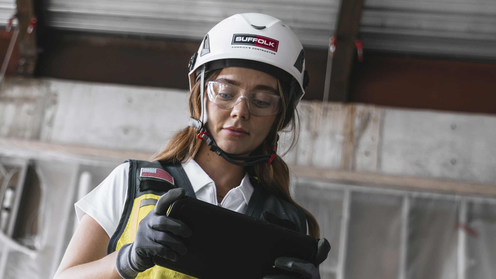 A construction worker wearing a white helmet, safety glasses, gloves, and a reflective vest looks down at a digital tablet. She stands in an industrial setting with metal and plastic sheeting in the background.