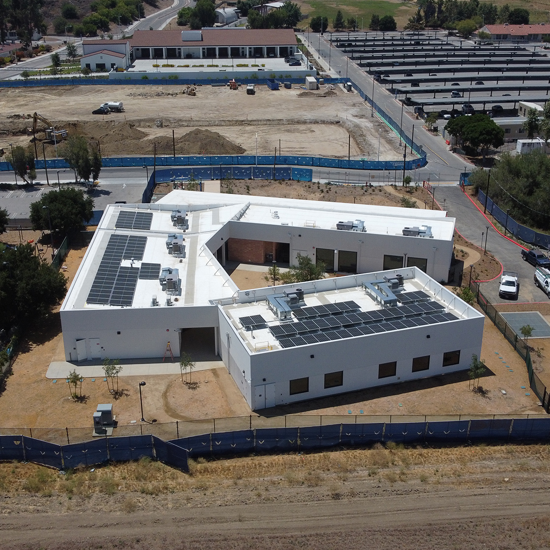 Aerial view of a modern white building with solar panels on the roof, surrounded by dirt and fencing, with construction and parking lots visible in the background.