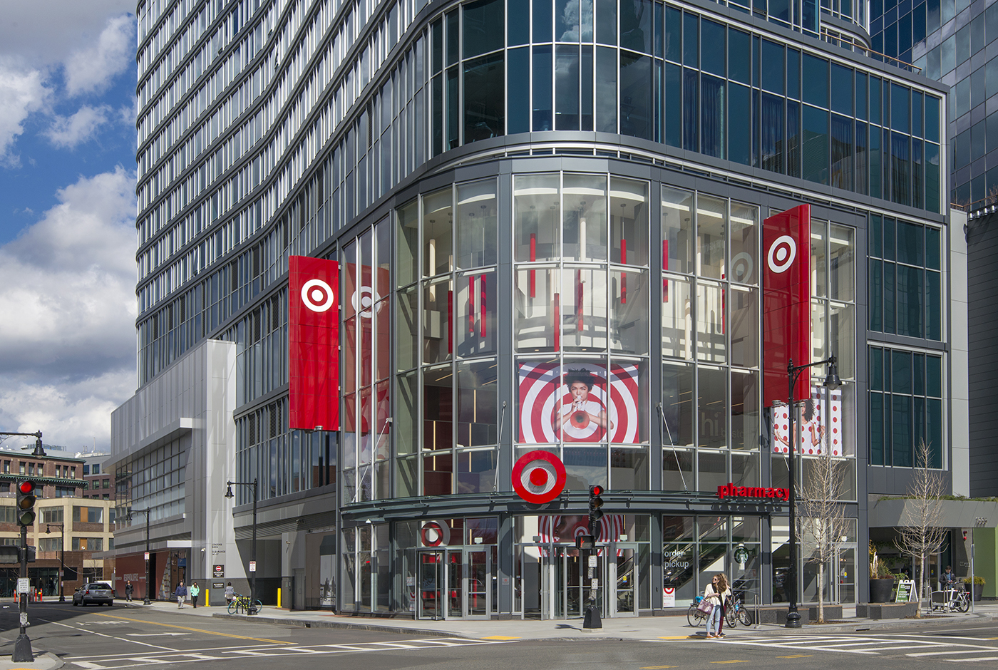 A modern glass building on a city street corner houses a Target store, with large red Target banners and logos visible on the facade and inside, and pedestrians walking nearby.