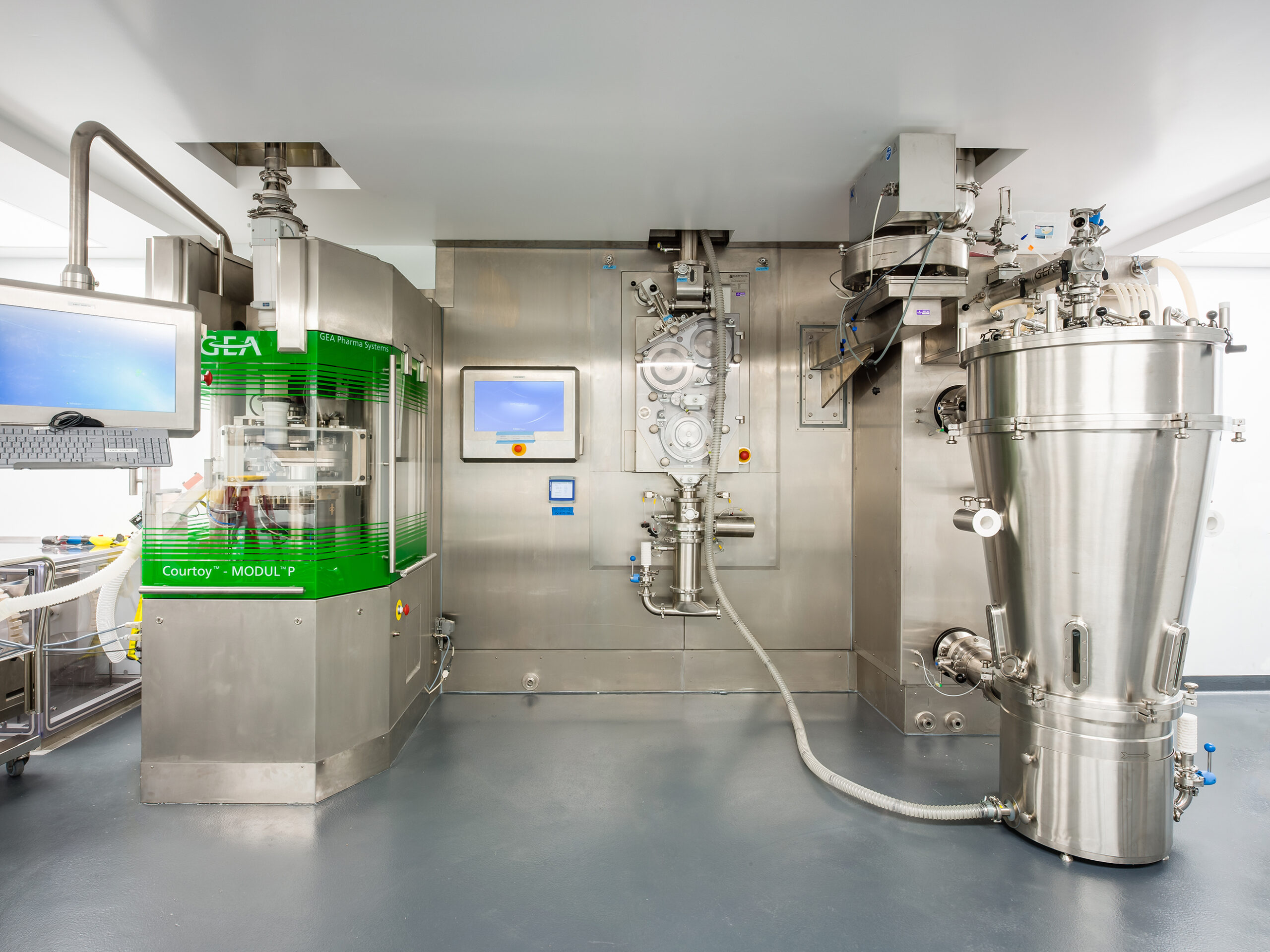 A modern pharmaceutical manufacturing room with stainless steel equipment, including a large machine on the left with green panels and a tall, cylindrical vessel on the right, both connected by pipes, under bright lighting.