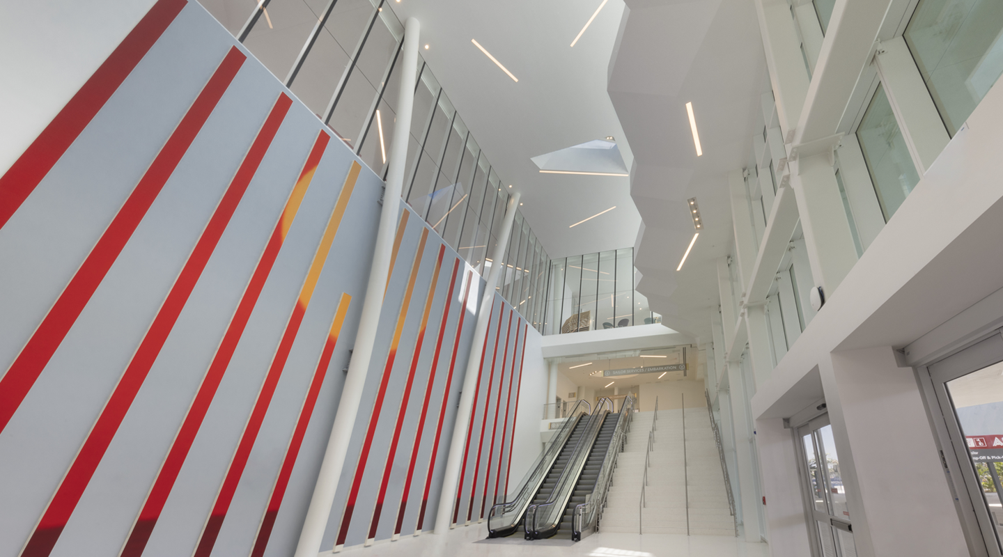 A modern, bright atrium with tall windows, red and yellow vertical wall accents, white floors, and a set of escalators and stairs leading to the upper level.