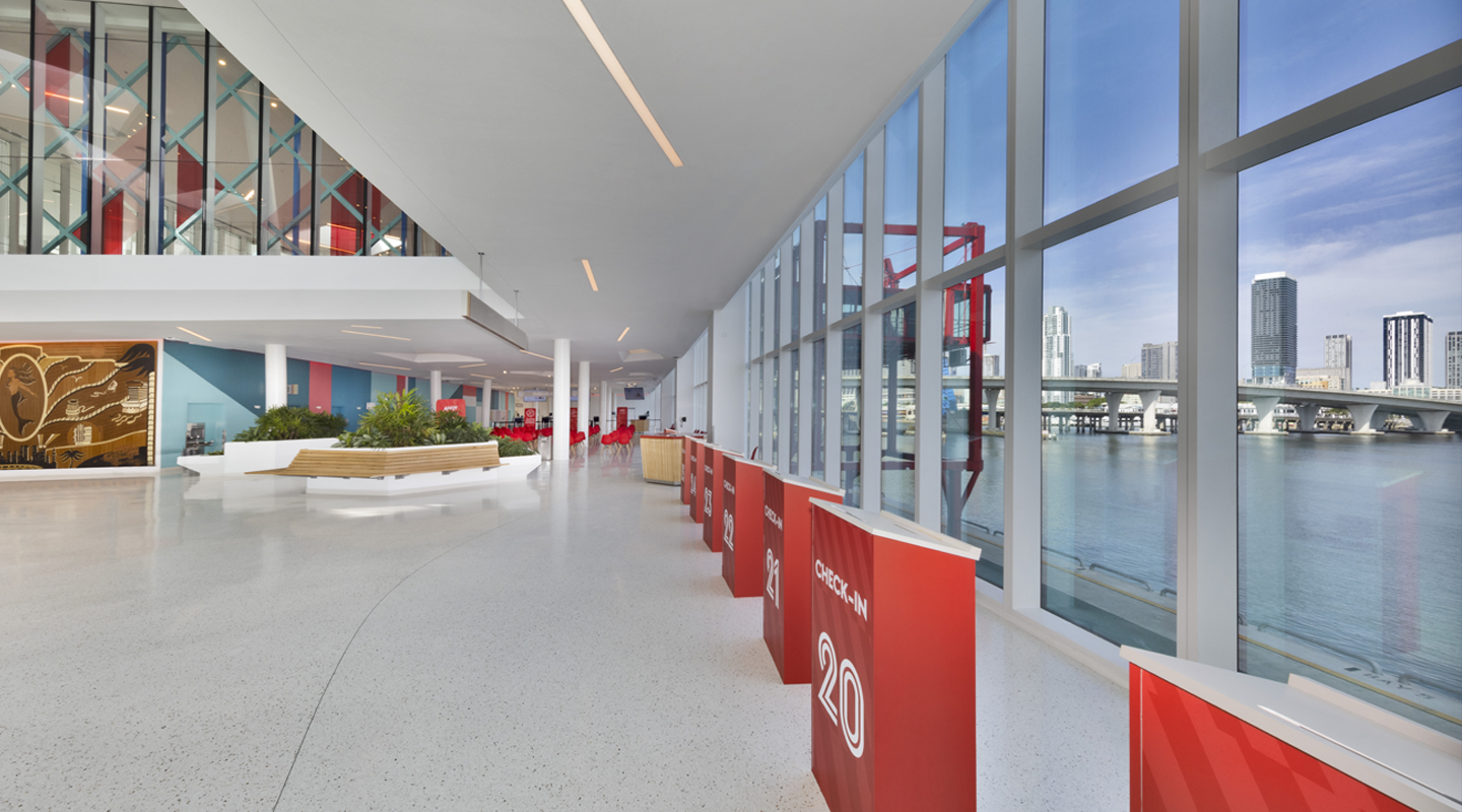 A modern, spacious interior with large floor-to-ceiling windows overlooking a city skyline and water. The area has red signs, indoor plants, and sleek white floors and walls, creating a bright, open atmosphere.