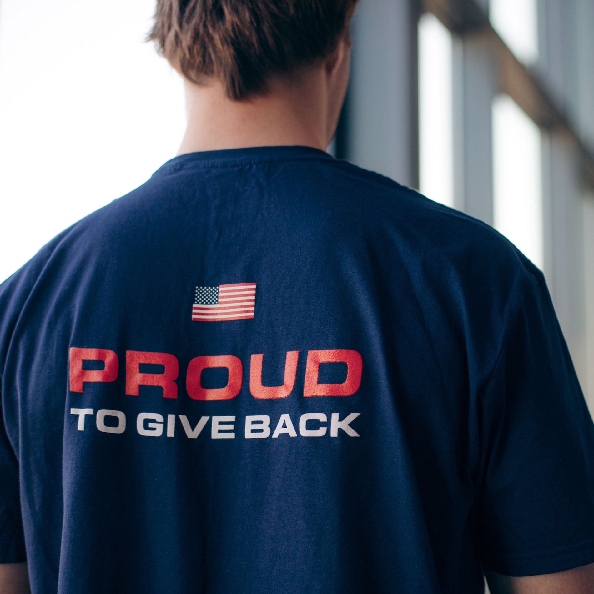 cropped zoomed in photo of the back of a persons tshirt with the words proud to give back and an american flag