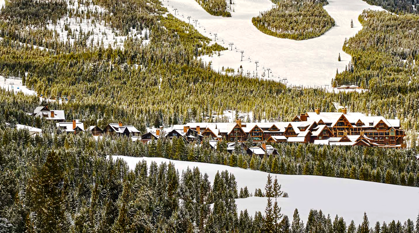 A snowy mountain landscape featuring a cluster of wooden lodges surrounded by dense pine forests, with ski slopes and ski lifts visible in the background.