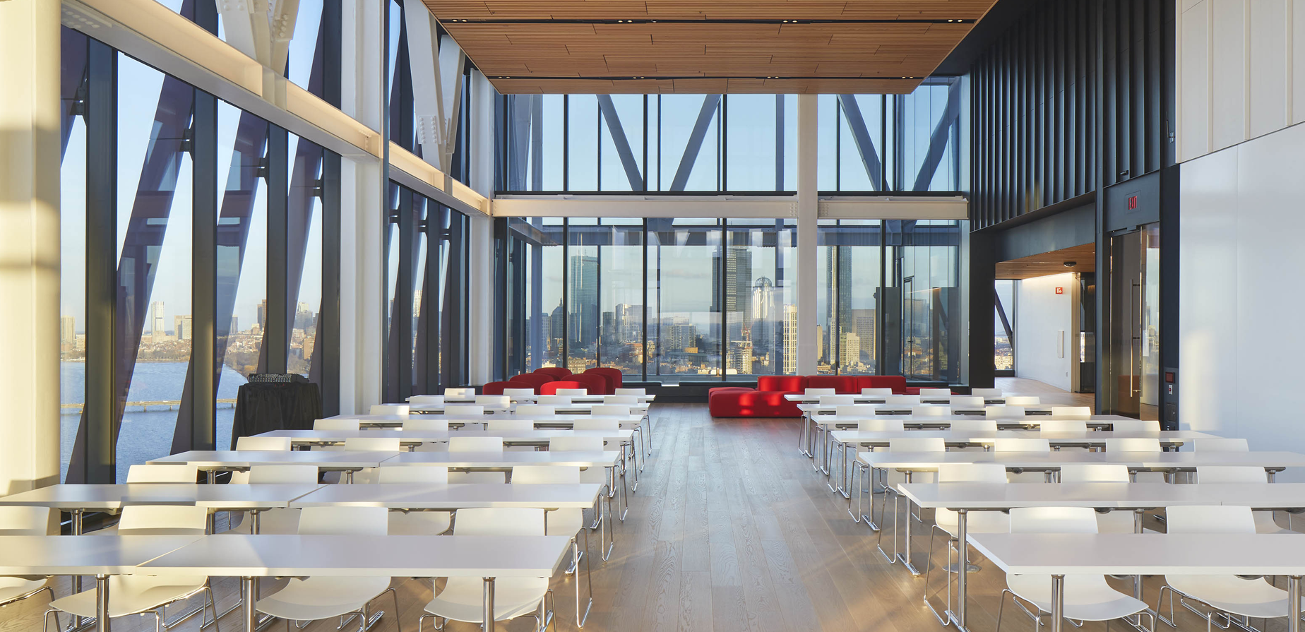 A modern classroom with large windows, white desks and chairs arranged in rows, light wood flooring, and red couches at the back, overlooking a cityscape and river through the glass walls.