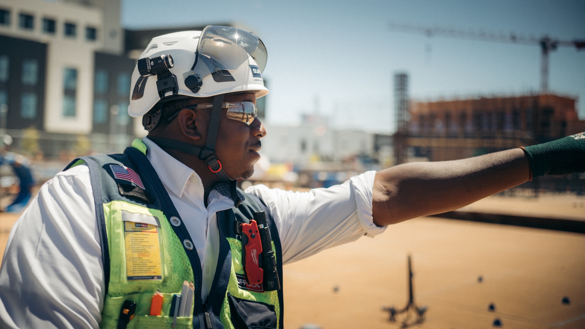 A construction worker wearing a safety helmet, reflective vest, and gloves points at something on a construction site, exploring potential careers amid unfinished buildings and cranes in the background.