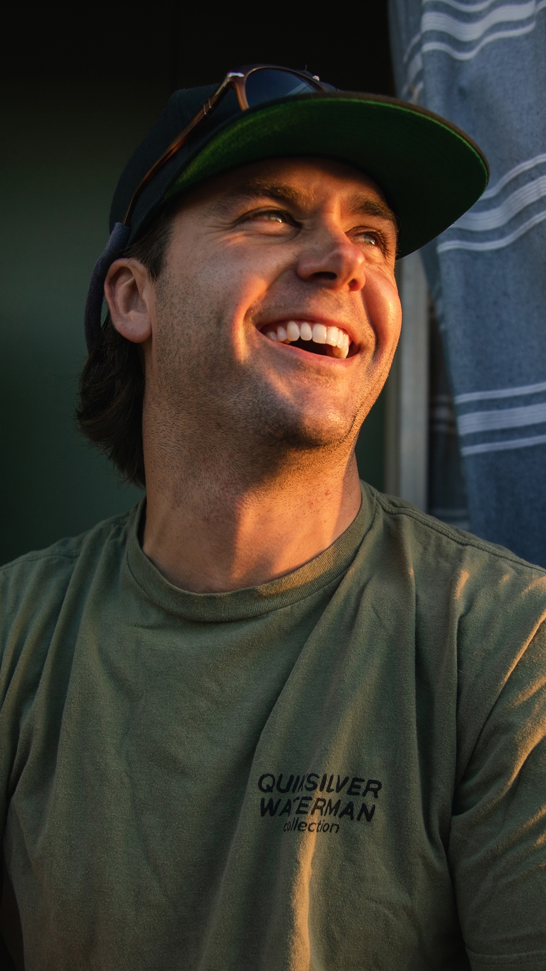 A young man wearing a green Quiksilver Waterman t-shirt and a dark cap smiles brightly, enjoying the benefits of warm sunlight on his face as he sits near a blue and white striped curtain.