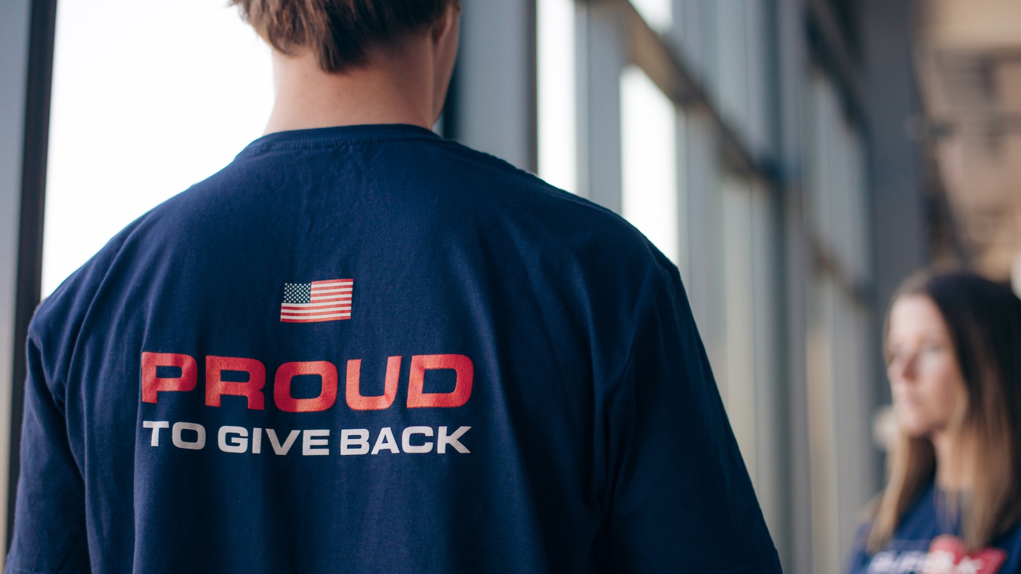 zoomed in photo of the back of a persons tshirt with the words proud to give back and an american flag