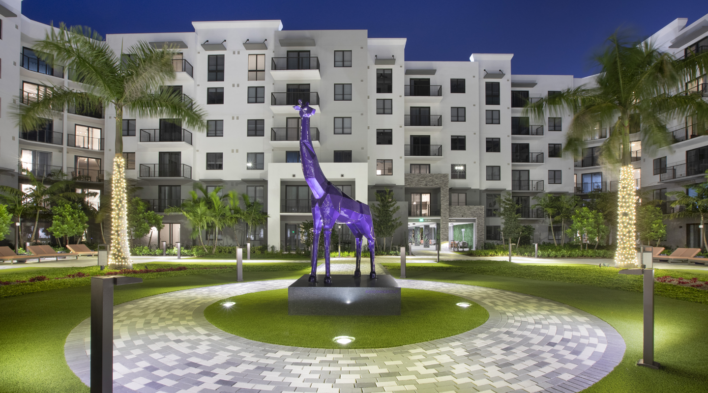 A modern apartment complex at night features a well-lit courtyard with a large purple giraffe sculpture in the center, surrounded by palm trees and circular pathways, with building balconies in the background.
