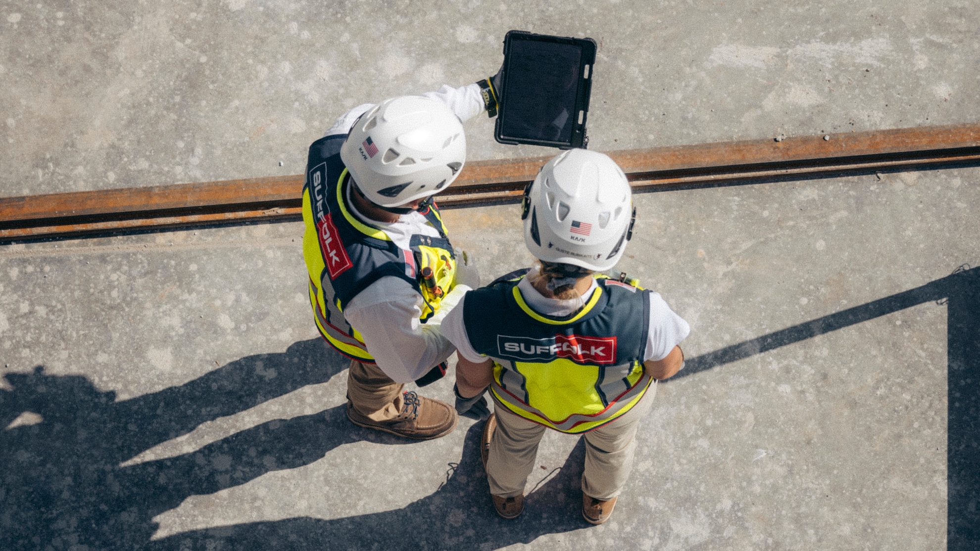 Two construction workers wearing hard hats and safety vests, each displaying Suffolk logos, stand on a concrete surface and look at a tablet together. The photo, taken from above, highlights teamwork and potential careers in construction.