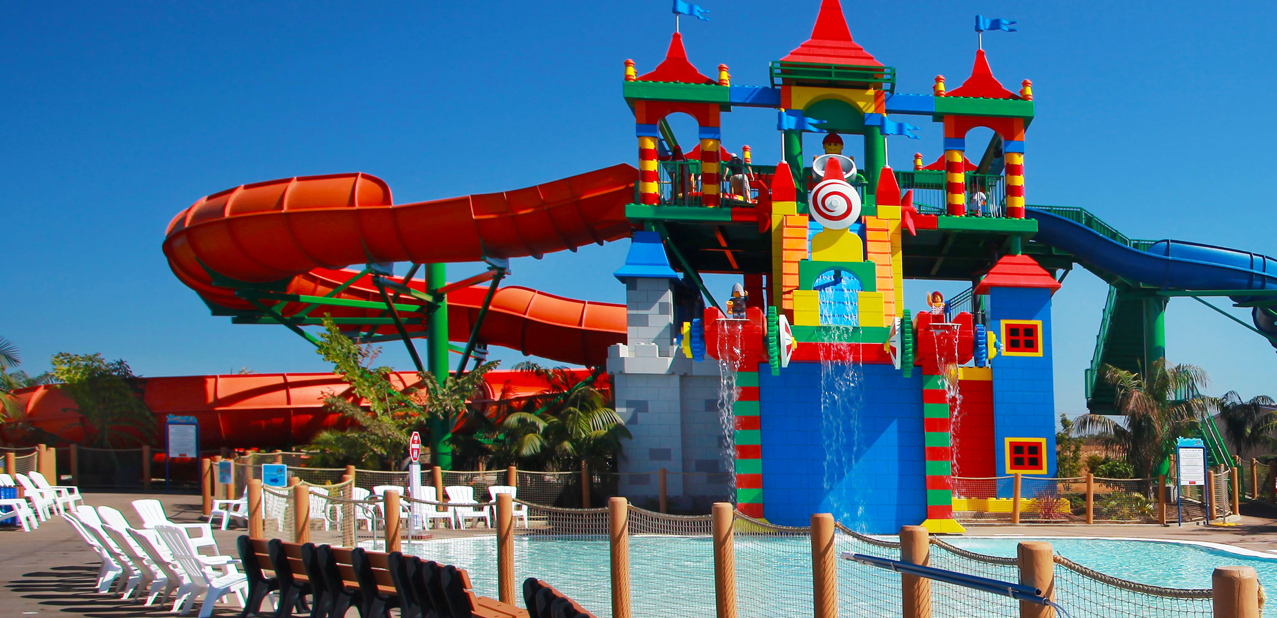 Colorful water playground with a castle structure, slides, and water features next to a shallow pool, surrounded by lounge chairs and palm trees under a clear blue sky.