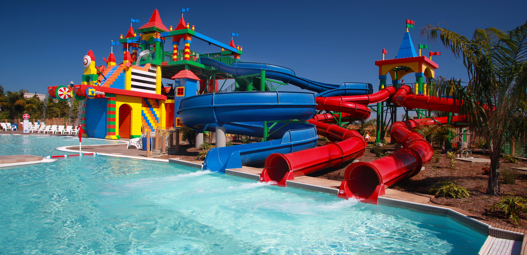 A colorful water park with twisting blue and red slides leading into a pool, next to a castle-themed play structure and surrounded by palm trees under a clear blue sky.