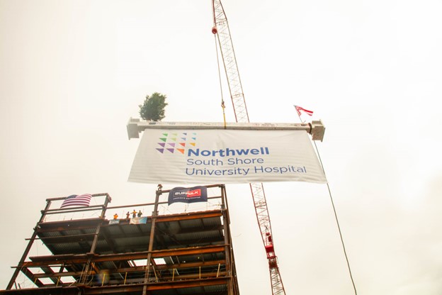 A crane lifts a steel beam with a small tree and a large banner that reads “Northwell South Shore University Hospital” at a construction site, with flags and workers visible on the building structure below.