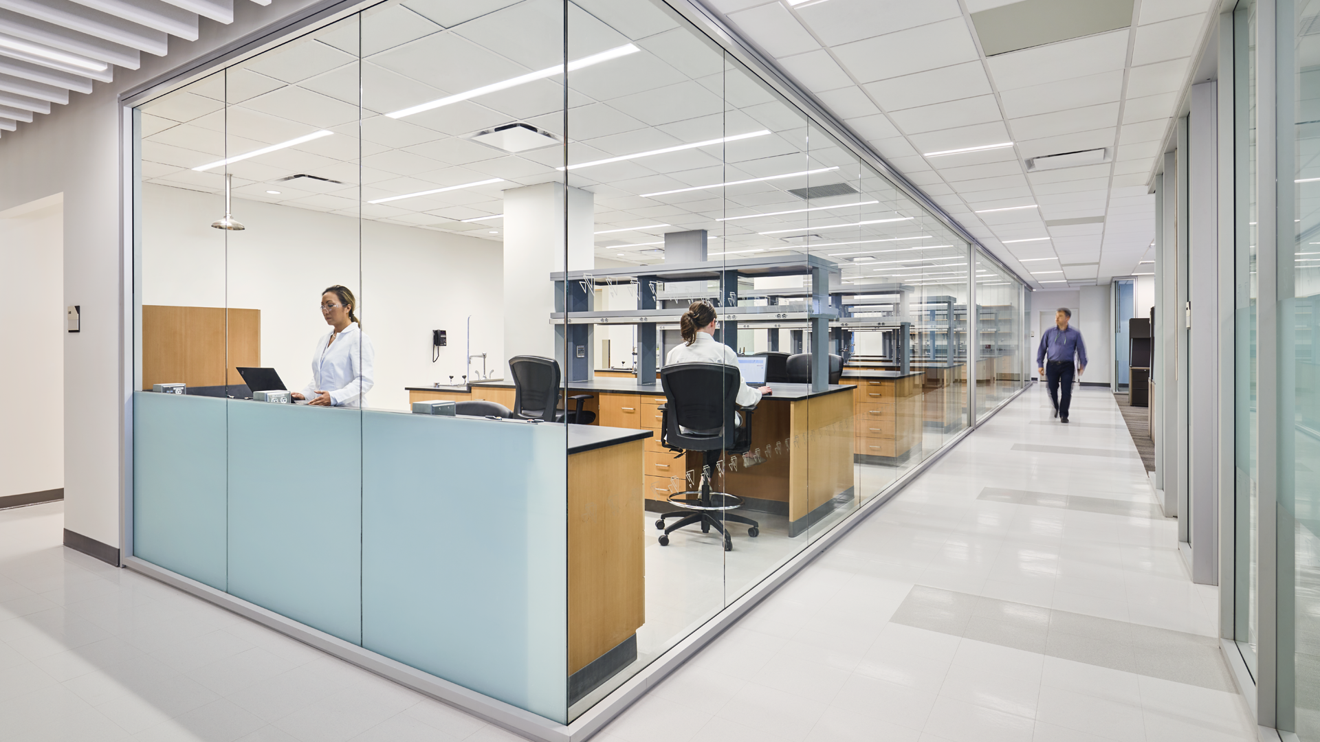 A modern laboratory with glass walls shows two people in lab coats working inside and one person walking by in the hallway outside. The space is clean, bright, and equipped with workstations and lab equipment.