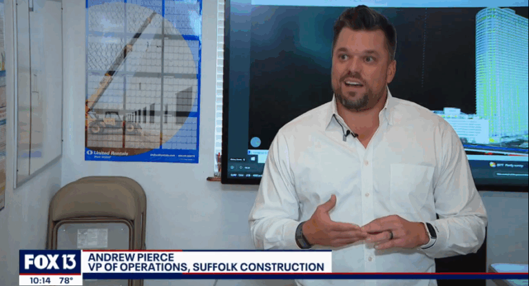 A man in a white shirt speaks while standing in an office with construction plans on the wall. A news banner at the bottom reads FOX 13 and identifies him as Andrew Pierce, VP of Operations, Suffolk Construction.