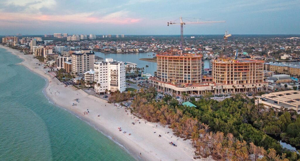 Aerial view of a sandy beach lined with hotels and buildings under construction, bordered by light blue ocean water and coastal vegetation, with a cityscape visible in the background.