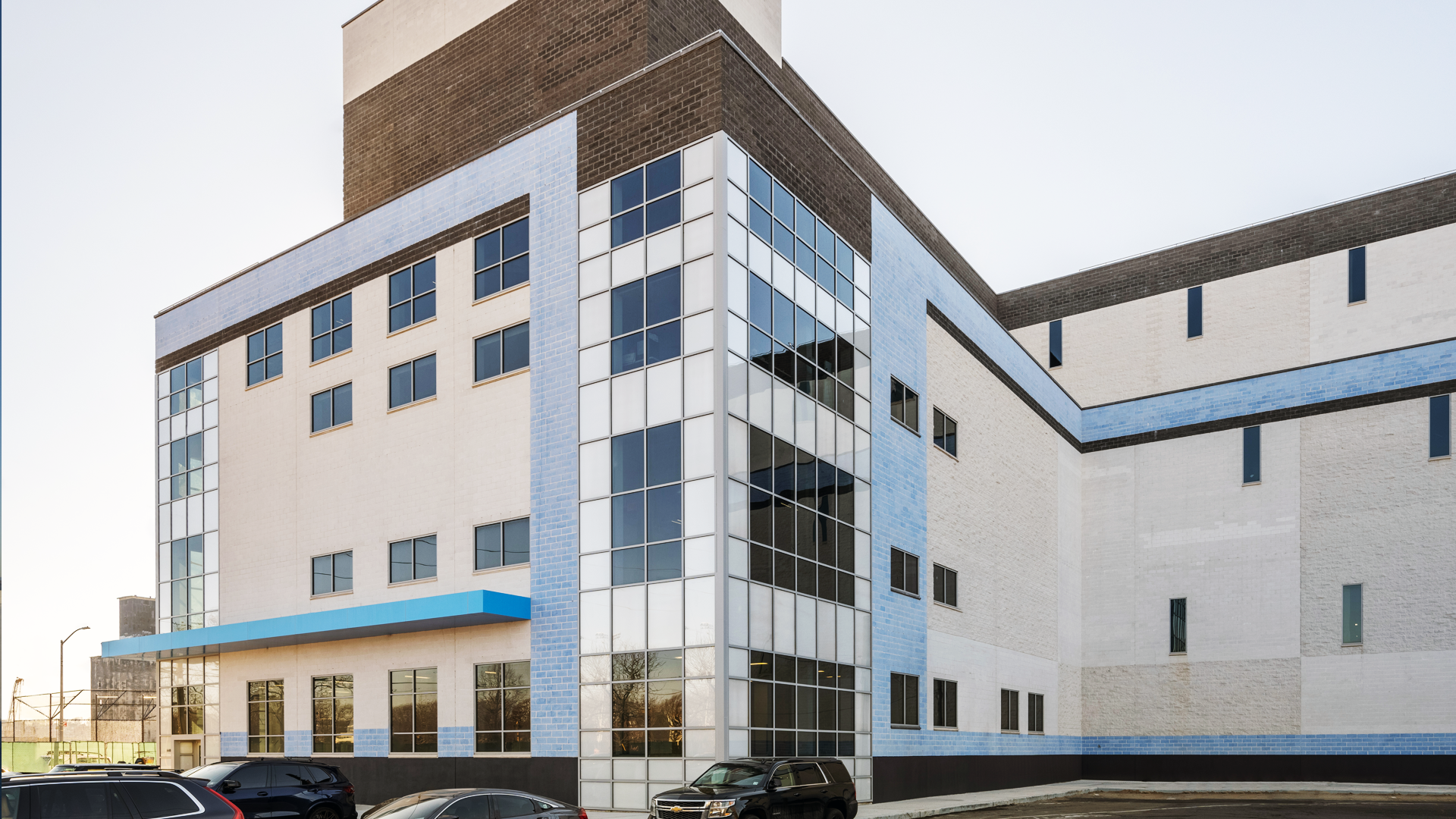 Modern commercial building with large glass windows, white and light blue exterior accents, and parked cars in front. The sky is clear and the building has multiple stories and geometric lines.