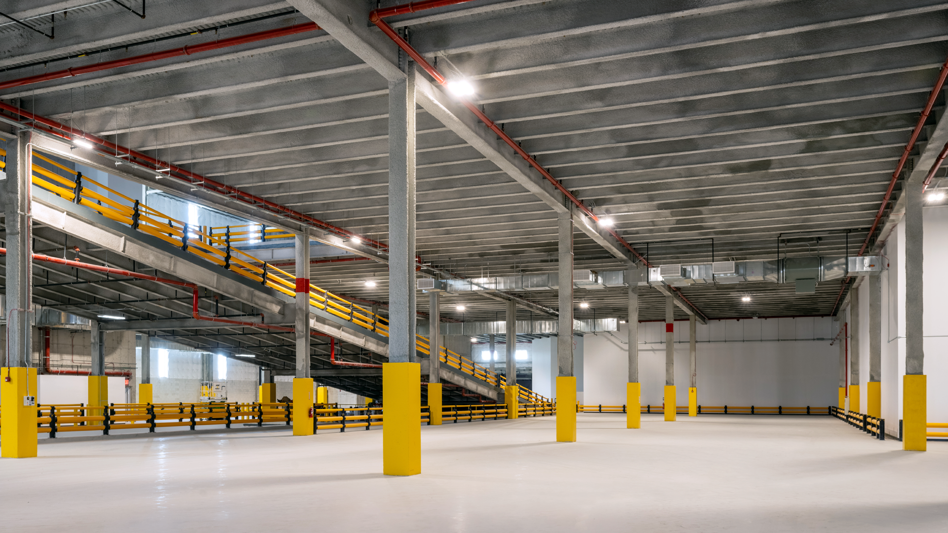 Spacious empty indoor parking garage with yellow safety barriers and pillars, concrete floor, and a ramp leading to an upper level. Overhead lights illuminate the clean, industrial space.