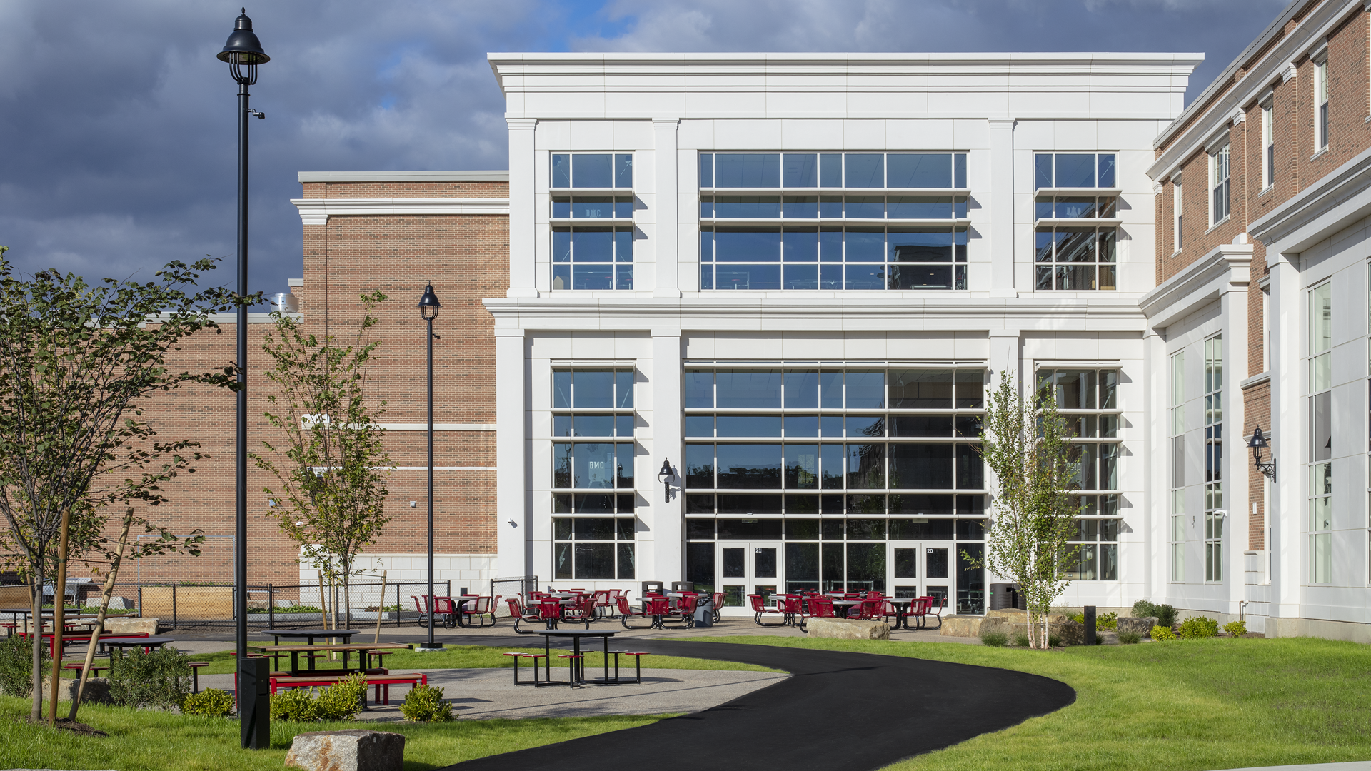 A modern school or university building with large glass windows, surrounded by green lawns and small trees. Red tables and chairs are arranged on the paved patio area under a partly cloudy sky.