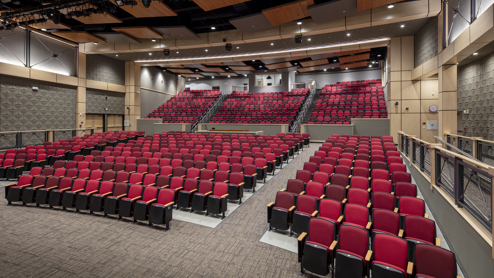 A modern auditorium with rows of empty red seats, gray walls, and a stage at the front. The seating is split into main floor and balcony sections, with bright, even overhead lighting.
