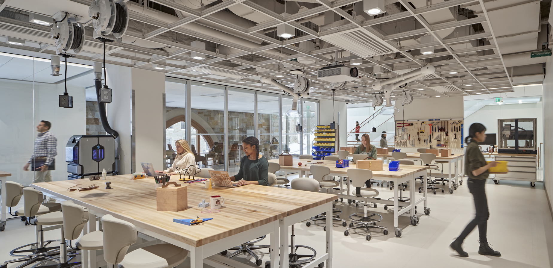 A spacious, modern makerspace with large wooden worktables, stools, and various tools. Several people are working or walking in the bright, well-lit room with glass walls and visible equipment storage.