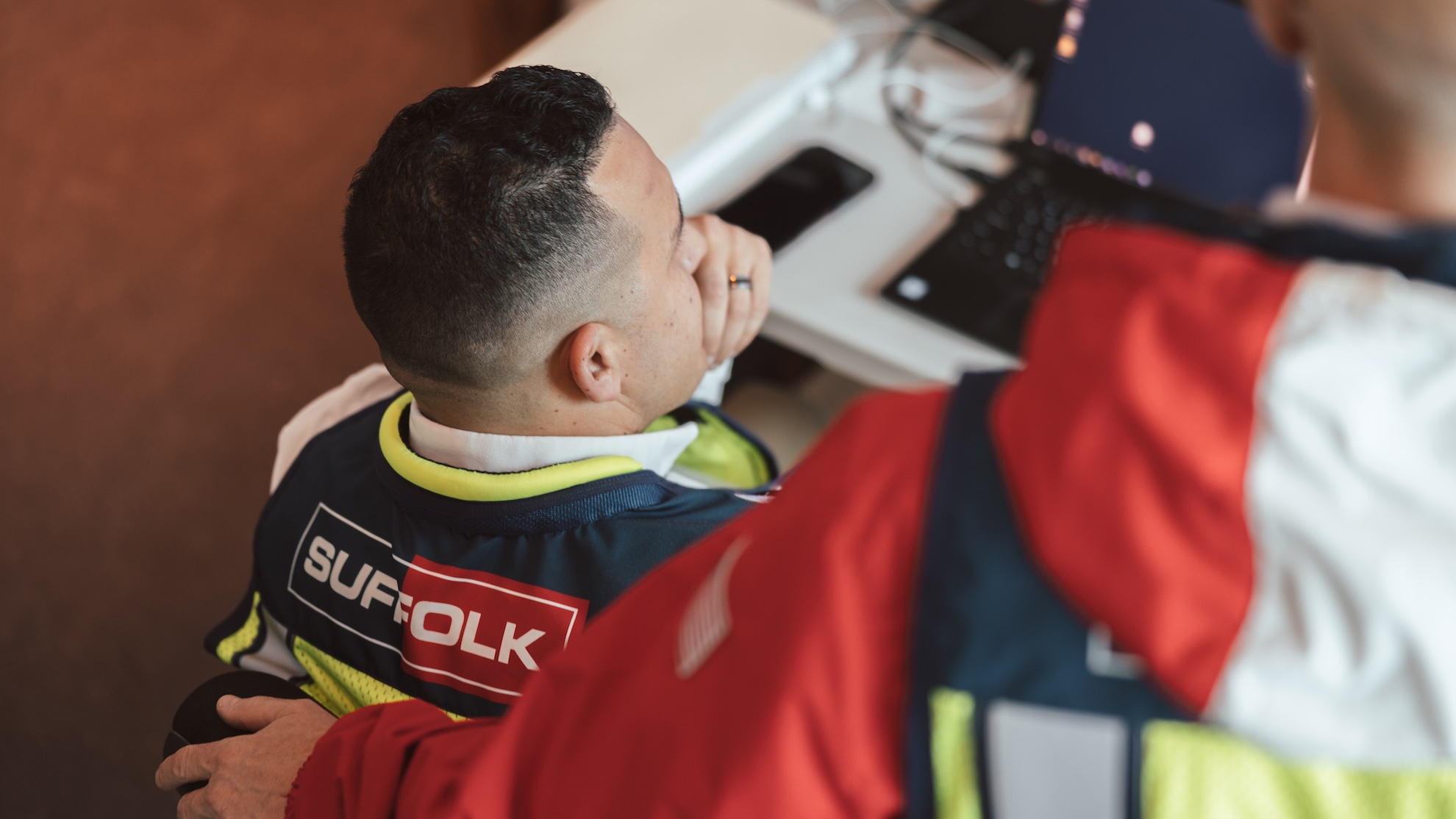 A man wearing a vest labeled Suffolk sits at a desk with a laptop and phone, while another person in a red jacket, possibly discussing careers, stands beside him, resting a hand on his shoulder.
