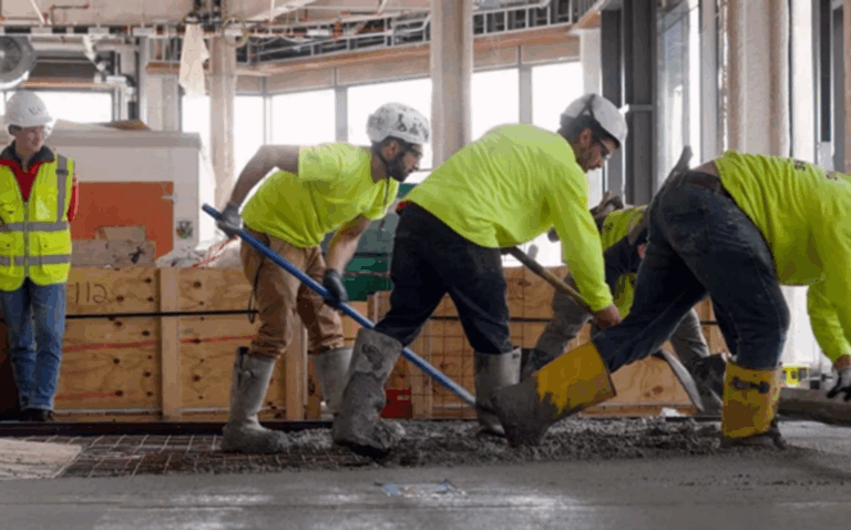 Four construction workers in safety gear and boots are spreading concrete on a floor inside a building under construction; a supervisor in a safety vest observes in the background.