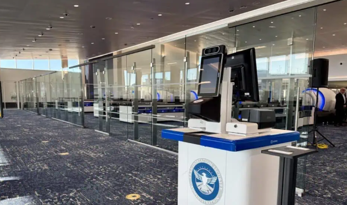 An empty airport security checkpoint with modern screening kiosks and glass barriers, featuring blue carpeting and bright lighting. No passengers or staff are present.