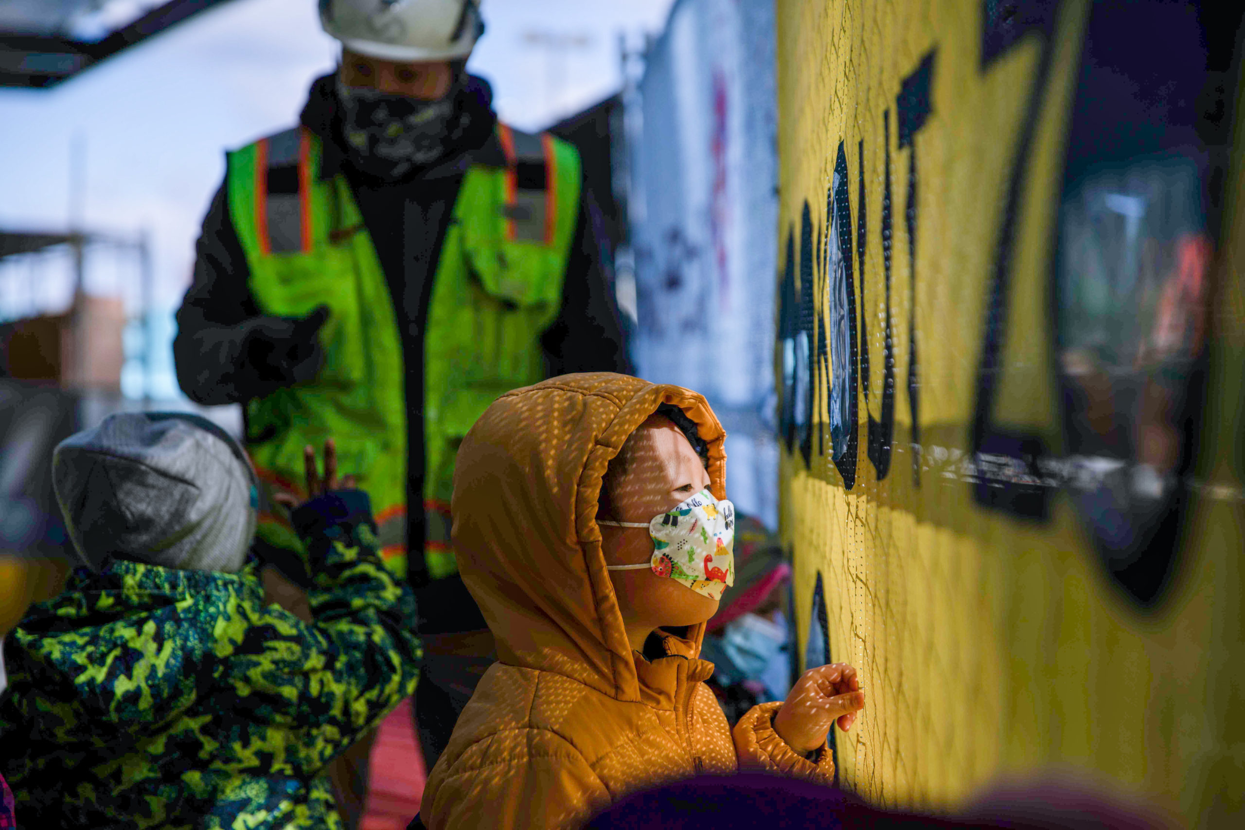 A child in a yellow coat and floral mask touches a yellow wall with black letters, while a construction worker and other children are nearby at a building site.