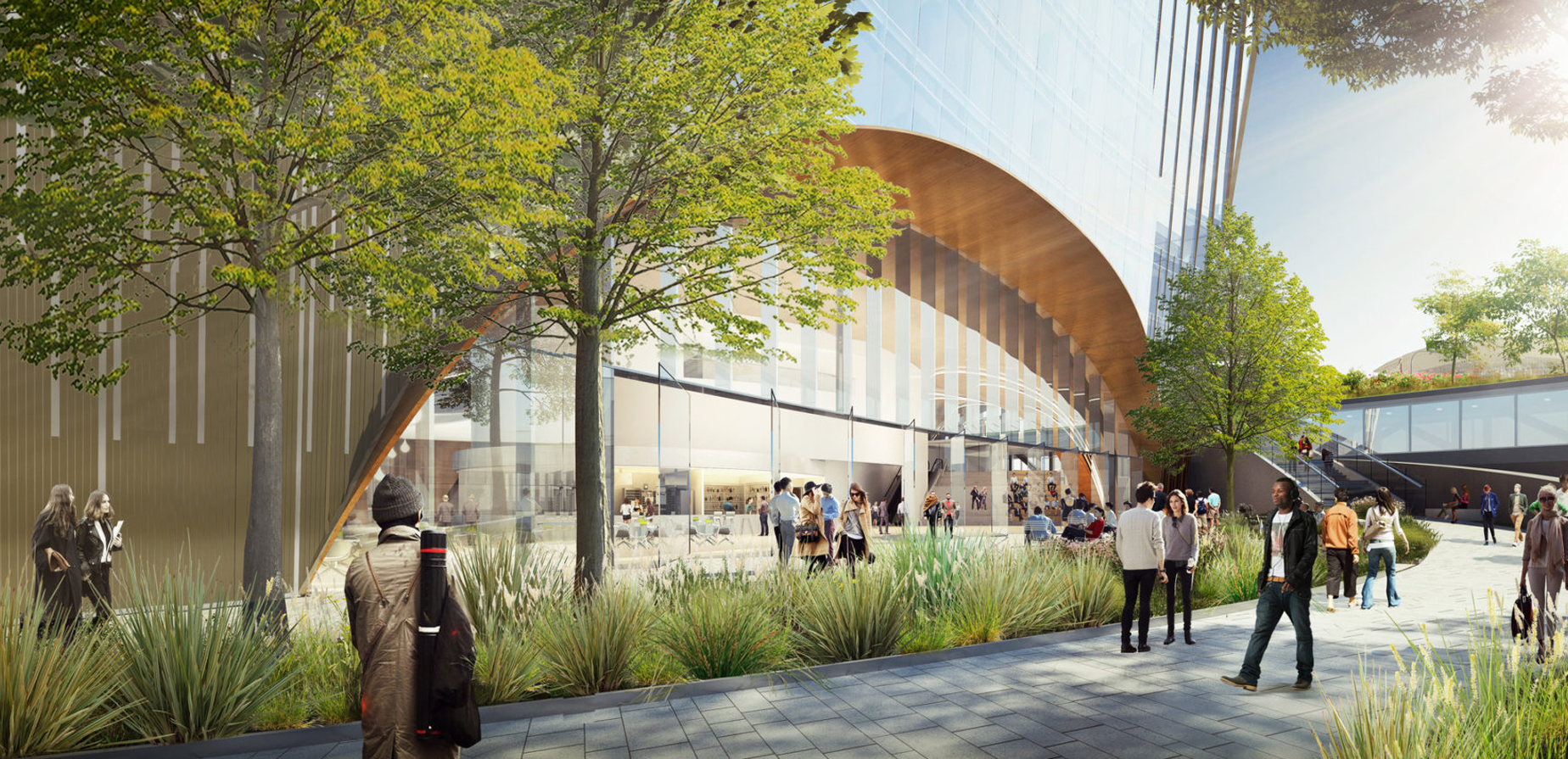 People walk and gather in a modern outdoor plaza with trees, greenery, and a glass building featuring large arched windows. Sunlight brightens the open space, and some people sit or converse near the building.