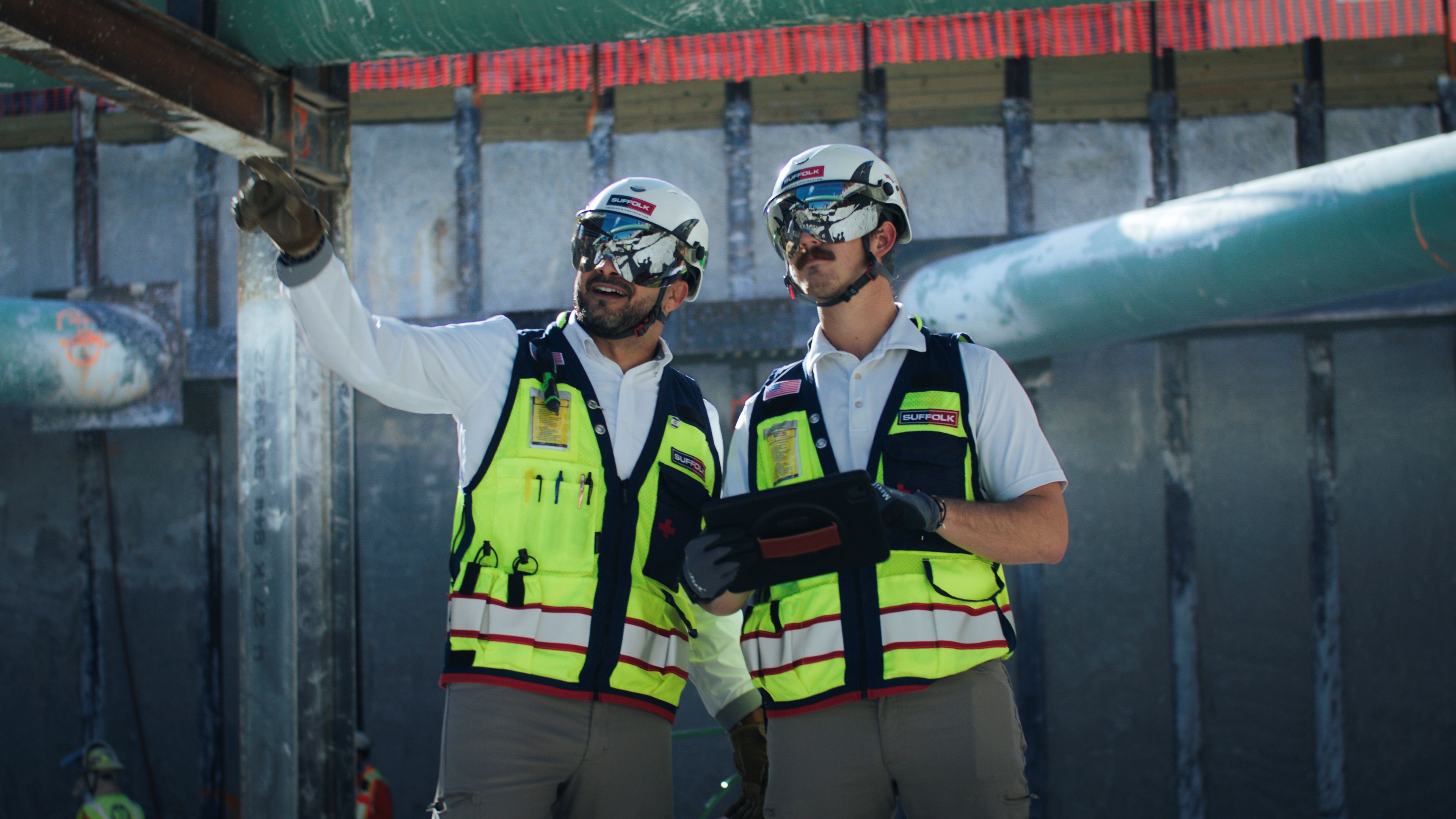 Two construction workers in safety vests, helmets, and sunglasses stand at a building site, showcasing rewarding careers. One points toward a structure while the other holds a tablet. Large pipes and a concrete wall are visible behind them.
