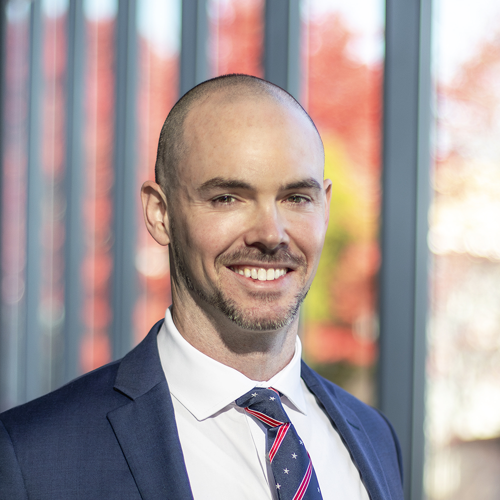 A smiling man with a bald head and short beard wears a dark blue suit, white shirt, and patterned tie, standing indoors with large windows and a blurred background of red foliage.