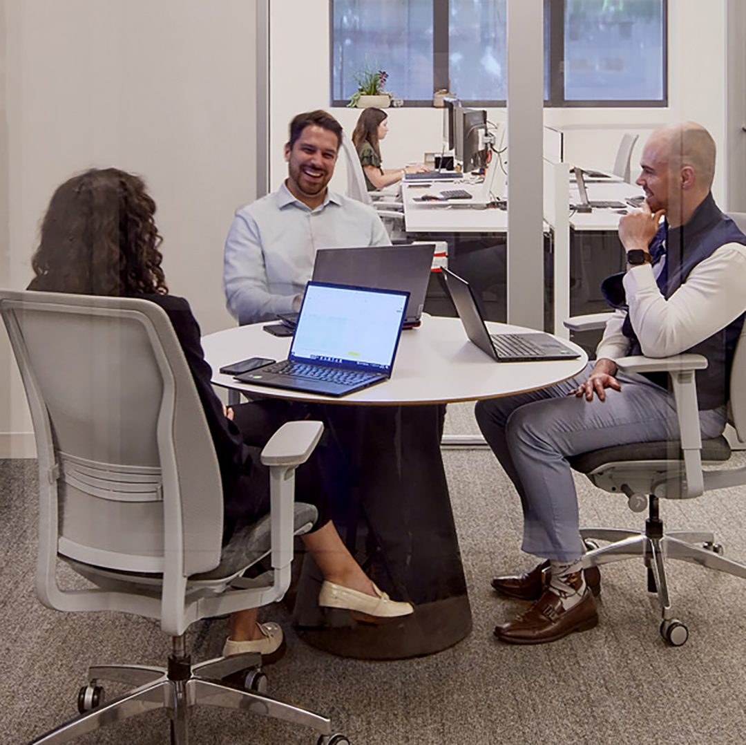 Three people sit around a round table in a modern office, each with laptops open, smiling and talking. More people work at desks in the background, separated by glass walls.