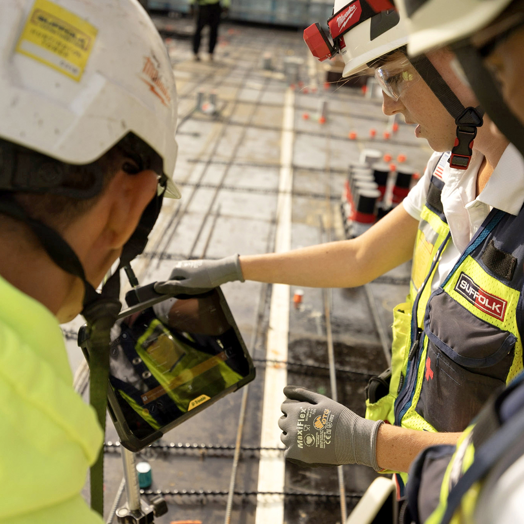 Two construction workers in safety gear and helmets review building plans on a digital tablet at a construction site, surrounded by rebar and materials.