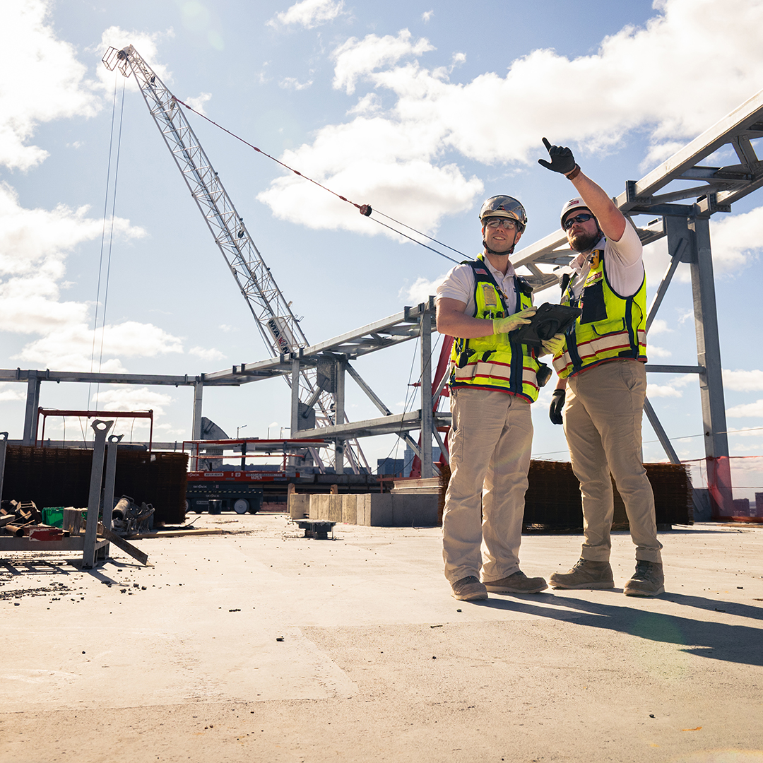 Two construction workers in safety vests and helmets stand on a construction site. One points ahead while the other smiles. A large crane and steel structure are visible under a partly cloudy sky.