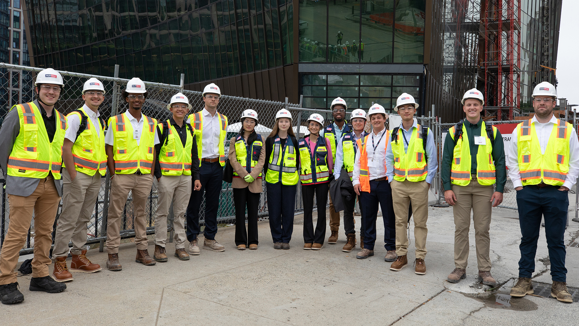 A group of fourteen people wearing hard hats, safety vests, and work boots stand side by side at a construction site with a building and fencing in the background.