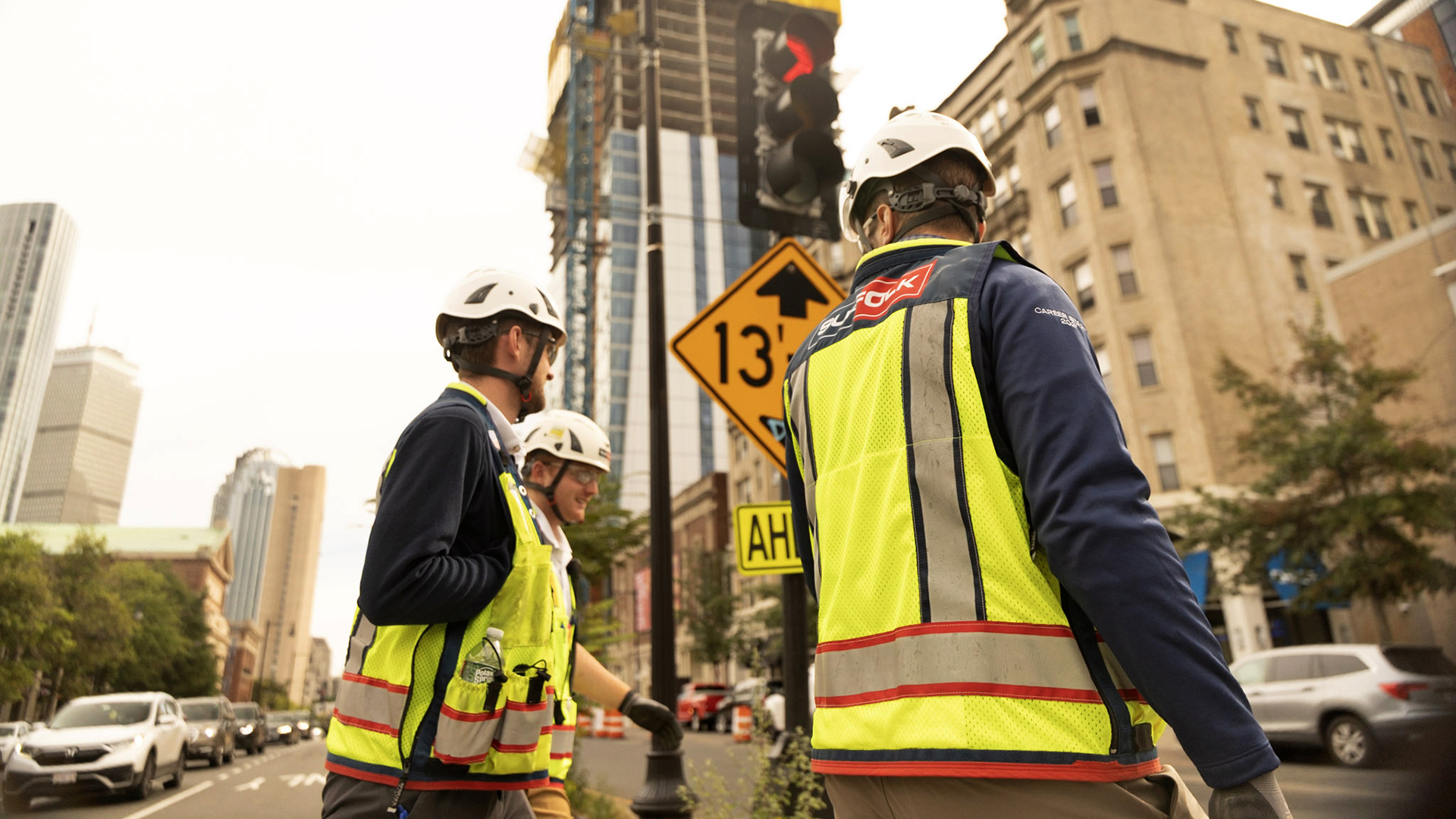 Three construction workers in safety vests and helmets stand on a city street near a construction site with tall buildings, a yellow height restriction sign, and a traffic light in the background.