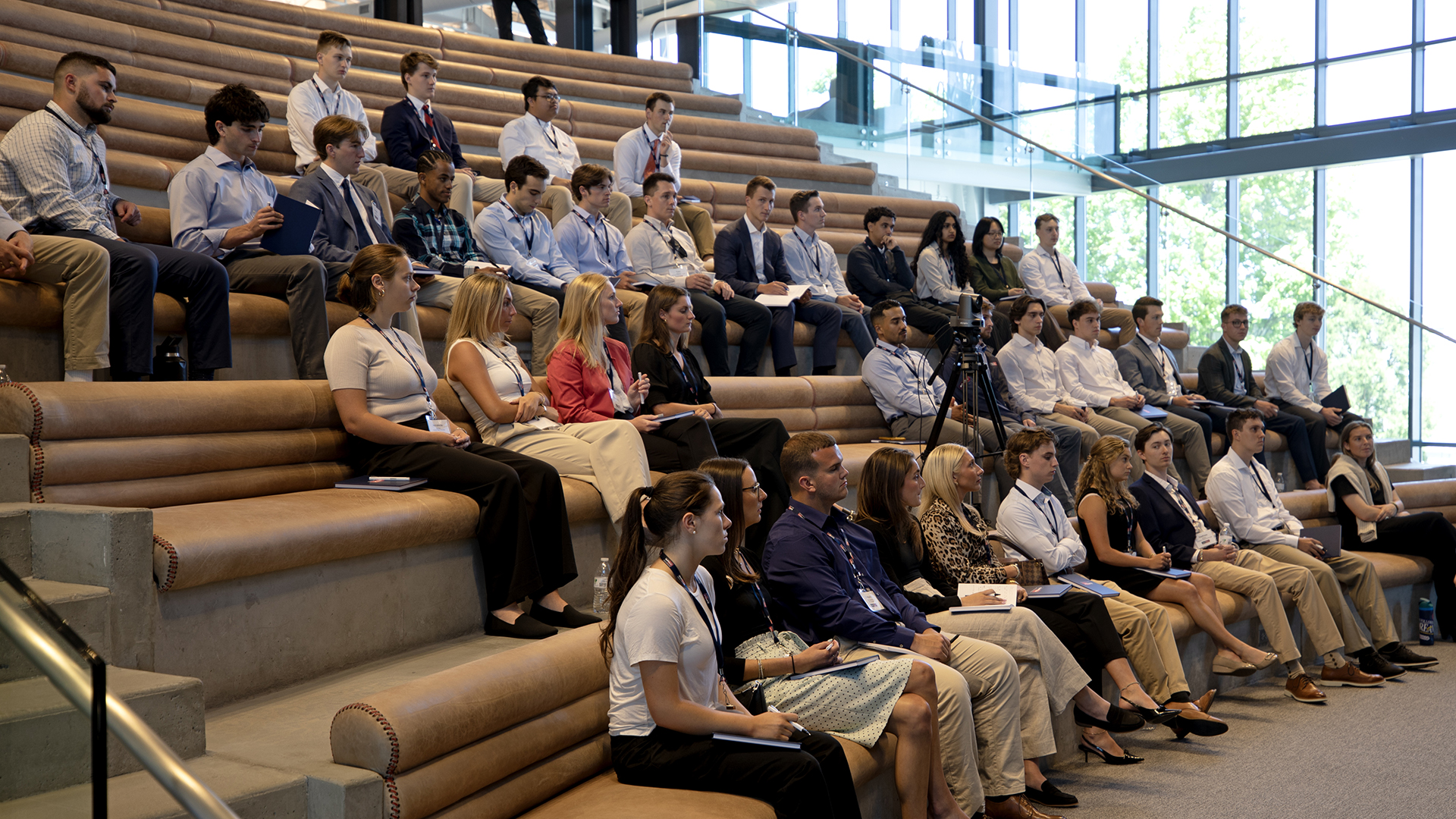 A group of people, mostly young adults, sit on tiered leather benches in a modern, sunlit auditorium, attentively watching a presentation or event. Some take notes, while a camera is set up in the middle aisle.