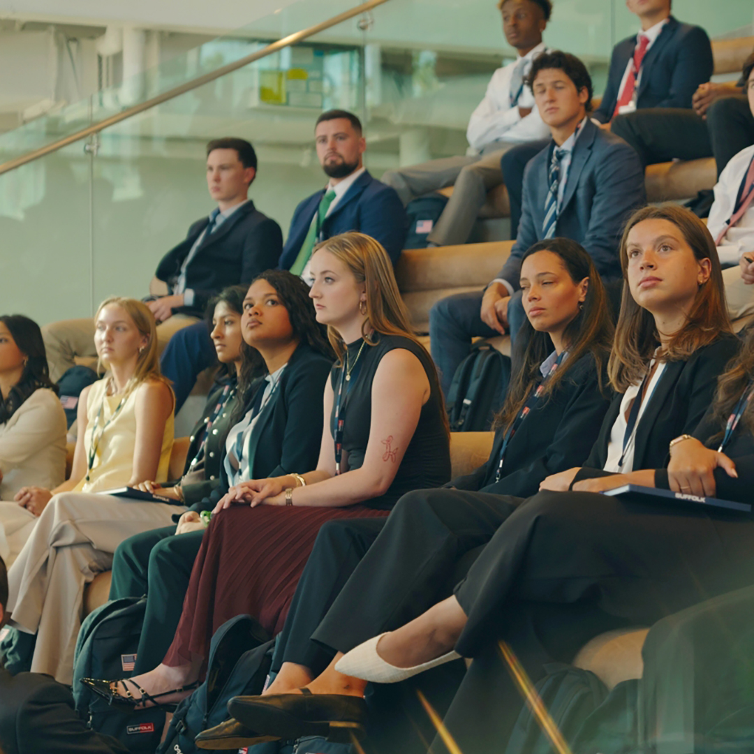 A group of professionally dressed young adults sit attentively on tiered seats in a modern, well-lit indoor space, possibly attending a conference or seminar.