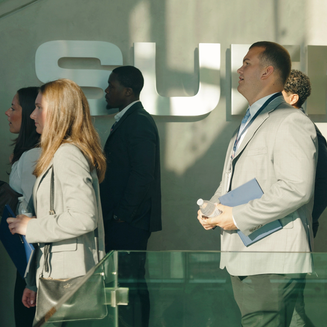 A group of professionally dressed people, some holding folders and a water bottle, stand indoors near a wall with large, partially visible letters in sunlight.