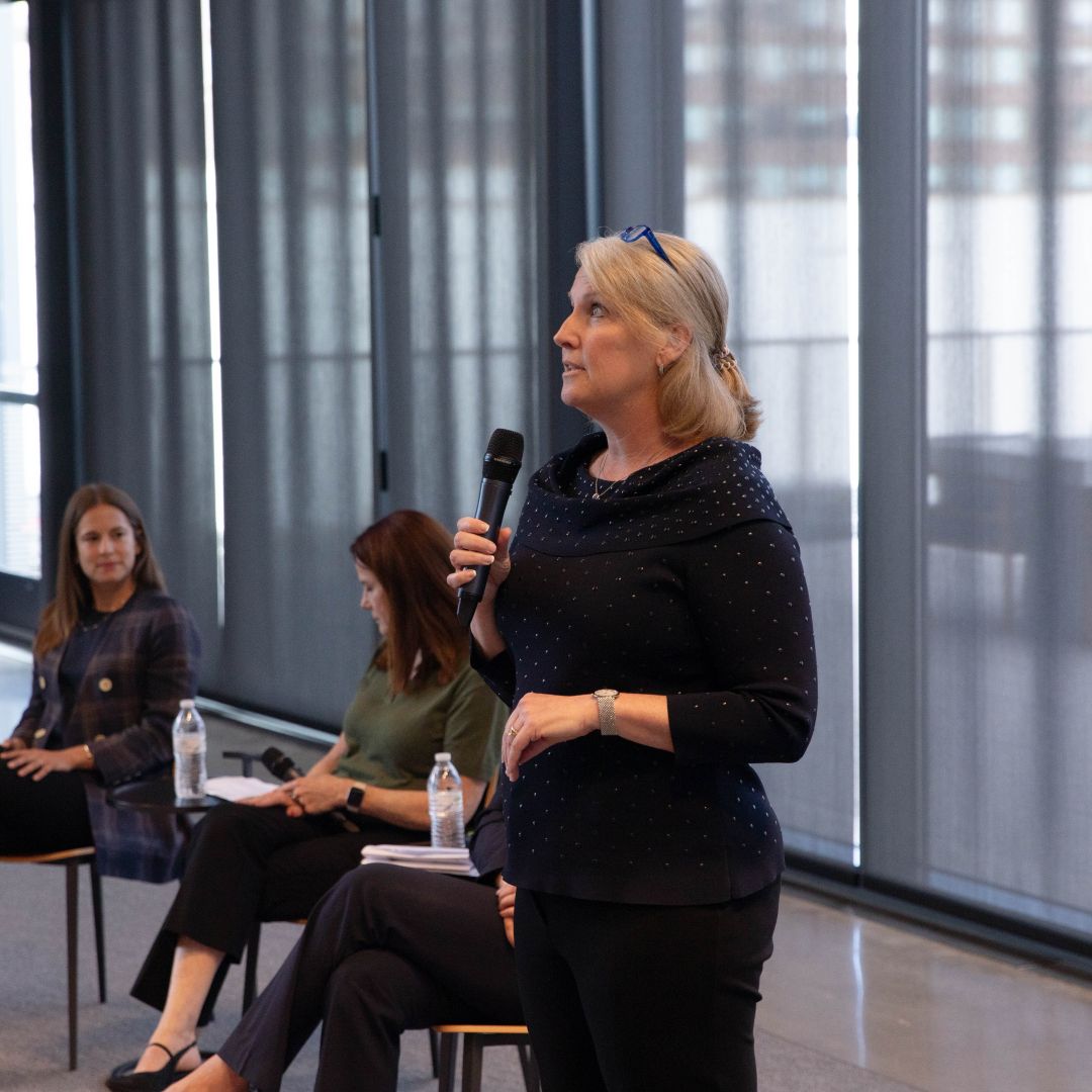 A woman stands and speaks into a microphone in a modern room during a careers discussion, while two seated women listen in the background. Large windows covered with sheer curtains let in natural light.