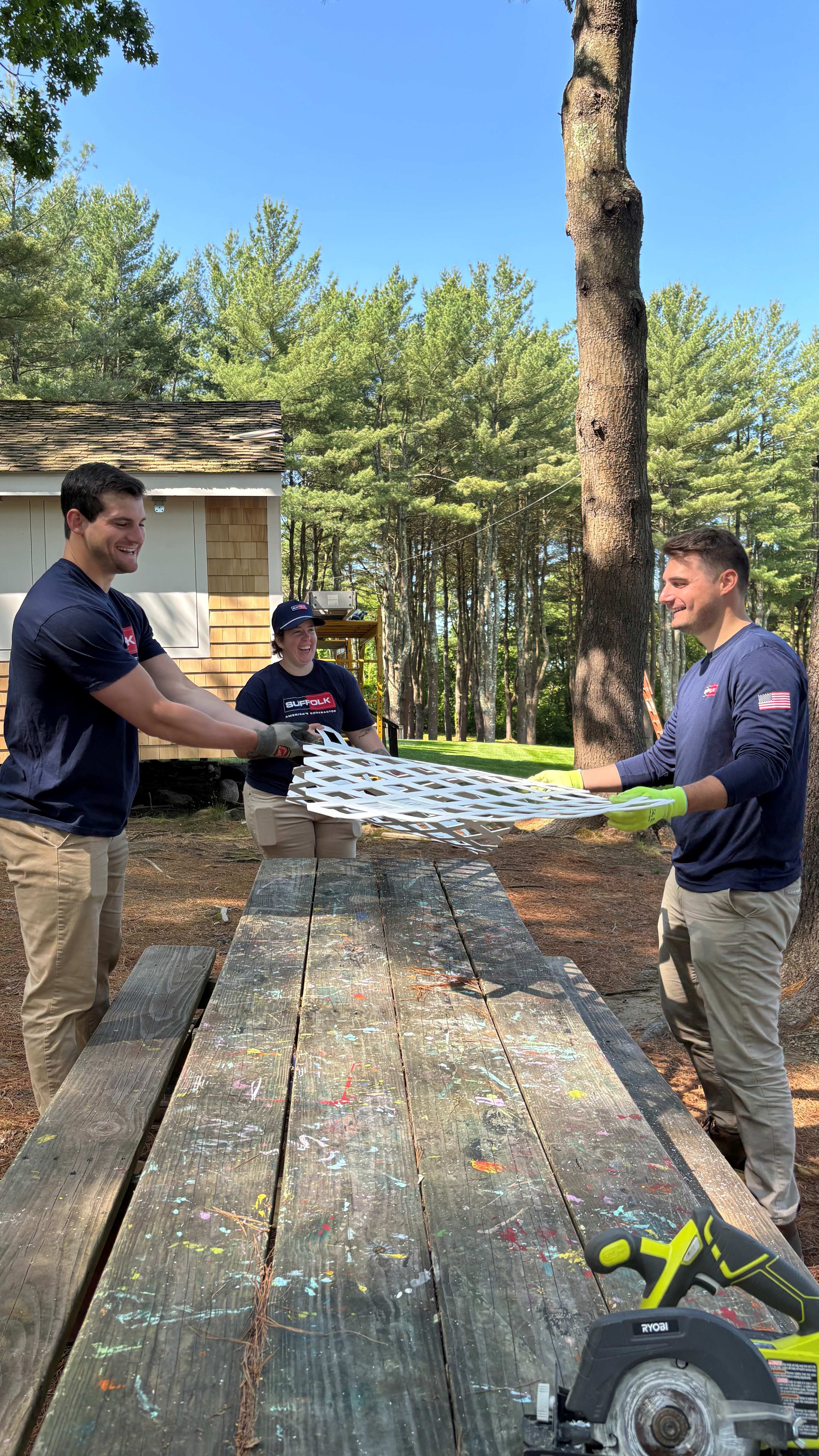 Three people outdoors, working together as a community—two stretching a metal sheet above a wooden picnic table while the third smiles nearby. They're surrounded by trees, with a cabin visible in the background.
