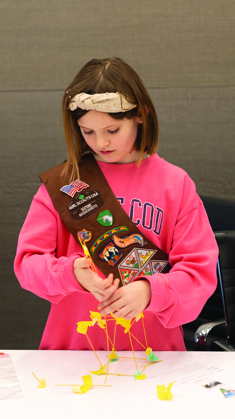 A Girl Scout wearing a brown sash and a pink sweatshirt builds a structure with yellow plastic pieces at a table, focused on her project. The plain background highlights her dedication to creativity and community.