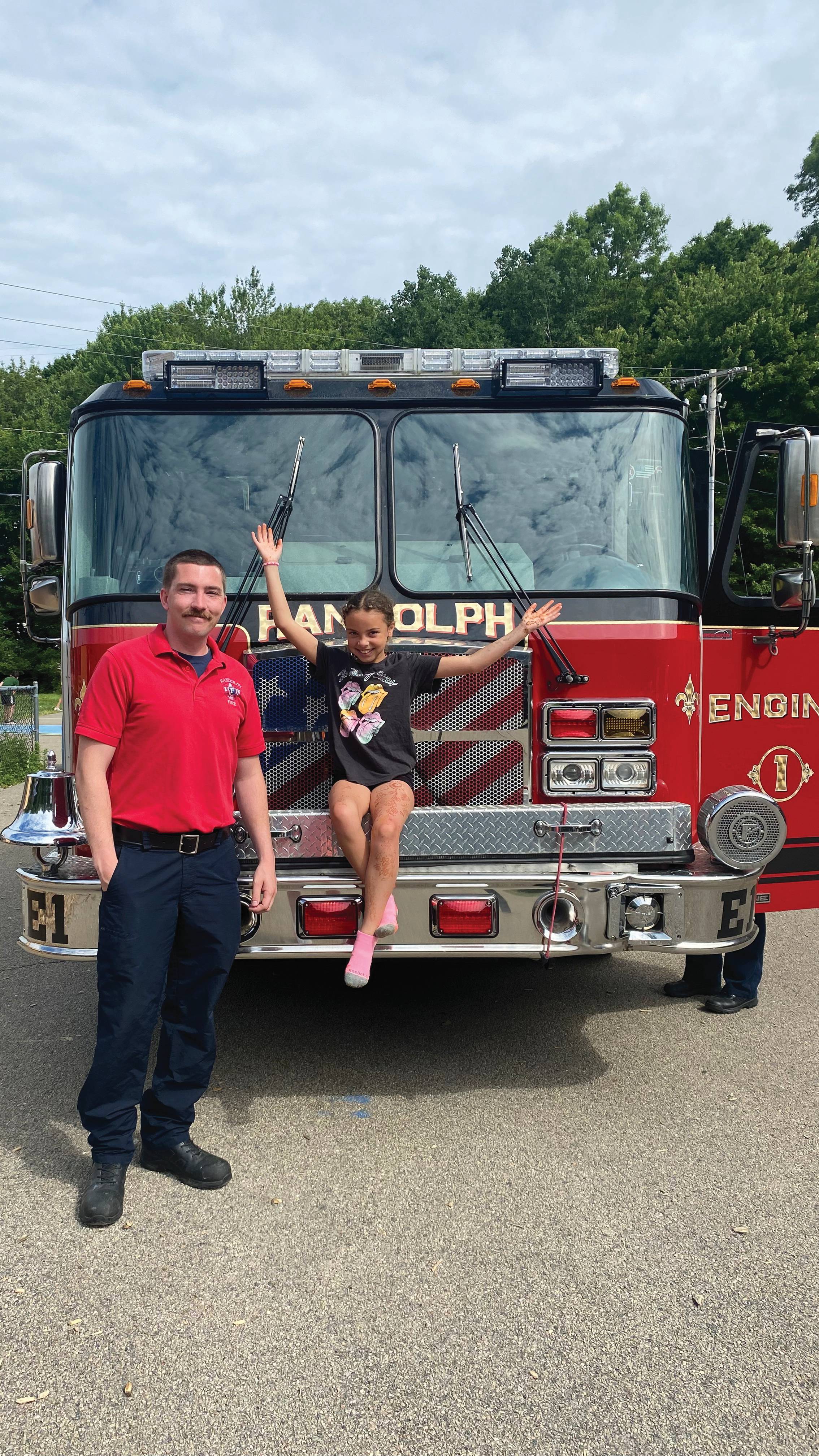 A community firefighter in uniform stands next to a smiling child sitting on the front bumper of a red fire engine with “RANDOLPH” on the windshield, outdoors on a sunny day.